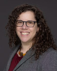 Susannah has dark curly hair and is smiling. She wears black-rimmed glasses and a charcoal grey suit with a chunky necklace.