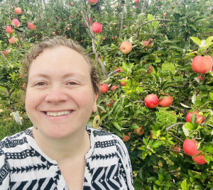 With her brown curly hair up, Beth smiles in an apple orchard. She is white and wears a white blouse with black triangles on it.