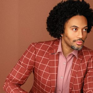 A stylish black man with full hair, salt and pepper stubble, and a dark anchor goatee looks off to the side. He wears a crisp button-up shirt that matches the color of his blazer's checkered stripes.