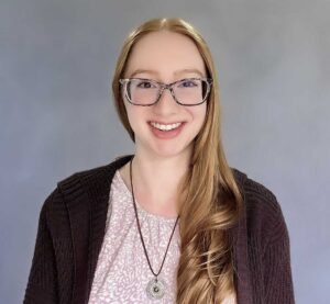 A young white woman with long, red hair smiles in front of a blue-gray portrait backdrop. She has purple tortoise glasses, a purple sweater, and a purple and white patterned shirt. 