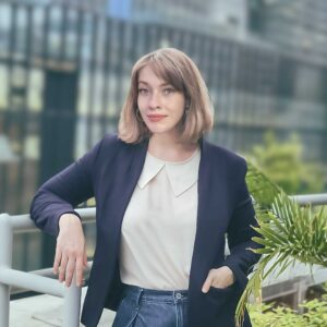 A white woman with chin-length brown hair leans on a rail with tall modern buildings behind her. She wears a navy blazer and a cream collared shirt.