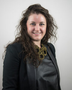 A white woman with dark wavy hair wears an and all black suit with chunky necklace smiles at the camera